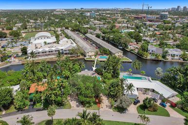 Aerial view of a sunny tropical waterfront neighborhood along a canal with palm trees, swimming pools, low‑rise apartments and city skyline in the distance; white arrow marking a waterfront property.