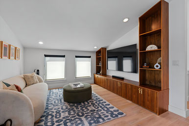Contemporary living room with a curved cream sofa, blue-gray patterned rug, round olive ottoman, low walnut built-in media cabinets and open shelving with a large flat-screen TV, two shaded windows, recessed lighting and light hardwood floors.