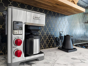 Modern kitchen stainless-steel coffee maker with red knobs and carafe on a marble countertop, black-and-gold tiled backsplash and matte black electric kettle under a wooden shelf.