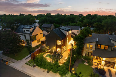 Aerial dusk view of a modern two-story home with black metal siding, illuminated backyard pool and string lights, solar panels on the roof, surrounded by contemporary houses and trees in a suburban neighborhood.