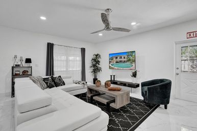 Bright modern living room with white sectional sofa, black patterned rug, wooden coffee table and green velvet armchair, wall-mounted TV showing a backyard pool, potted plant, overhead ceiling fan and exit sign by the door.