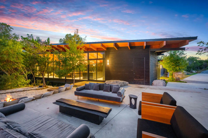 Modern wood-clad house with floor-to-ceiling windows and sloped roof, overlooking a furnished concrete patio with sofas, wood bench, fire pit and trees glowing in a colorful sunset.