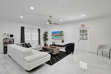 Bright modern living room with white sectional sofa, black accent chair and patterned rug, wood coffee table, wall-mounted TV showing a pool, ceiling fan, potted plant, marble tile floor and exit door — contemporary vacation rental interior.