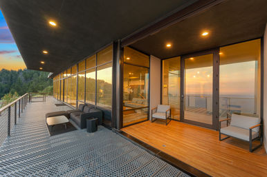 Modern glass-walled hillside home balcony with metal grated deck, black sectional sofa, outdoor chairs and warm interior lighting, overlooking forested coastline and ocean at sunset.