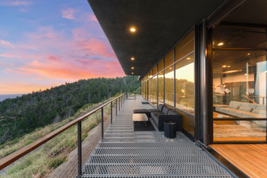 Modern hillside home balcony with metal grating and glass wall, outdoor lounge overlooking a forested valley at pink‑orange sunset