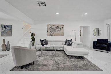 Bright, airy modern living room with white L-shaped sectional, navy accent pillows and velvet armchair, glass coffee table on a textured rug, abstract wall art, potted plant, marble floors and recessed lighting.