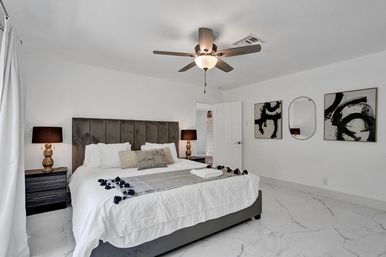 Bright, spa-like modern master bedroom with king bed and velvet headboard, ceiling fan, marble-look tile floor, bedside lamps and abstract wall art.
