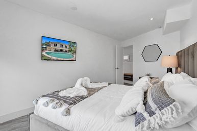 Bright, cozy modern vacation-rental bedroom with plush king bed, textured throw pillows and bedside lamp; wall-mounted TV showing a backyard pool and open doorway to hallway.