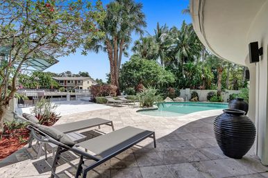 Sunny tropical waterfront backyard oasis with curved swimming pool, palm trees, tiled patio, lounge chairs, decorative urns and a private dock in the background