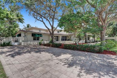 Sunlit two-story Mediterranean-style home with clay tile roof and green awnings, wide paved driveway and lush landscaped front yard with mature oak and palm trees under a clear blue sky.