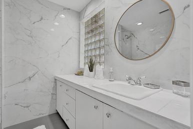 Bright spa-like modern bathroom with white marble walls and countertop, built-in sink and chrome faucet on a white vanity, round gold-framed mirror, glass-block window, and small potted plant.