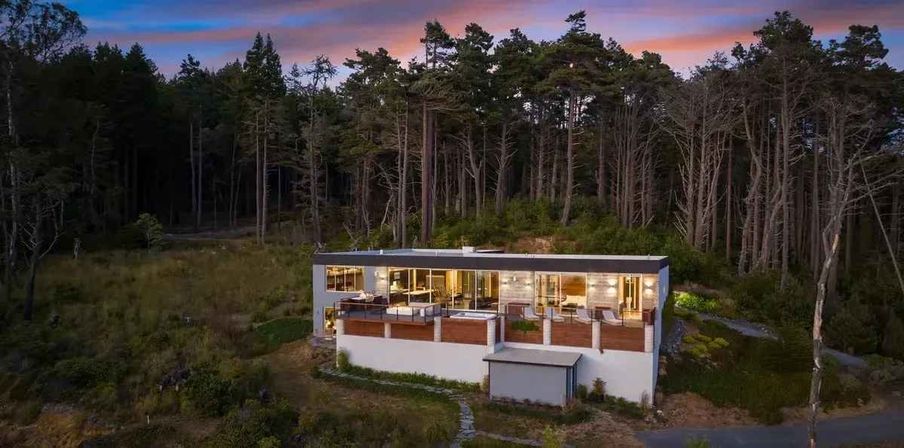 Contemporary glass-front hillside house with warm interior lights and wooden terraces, nestled at the edge of a tall pine forest under a pink-purple twilight sky — a cozy modern hideaway at dusk.