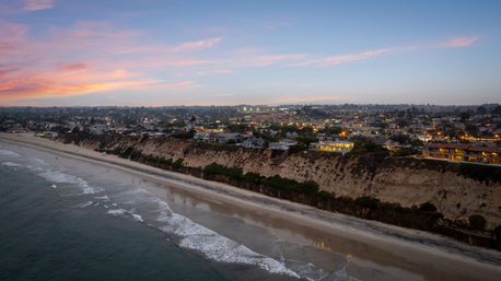 Sunset over a coastal bluff and sandy beach, gentle ocean waves lapping the shore with homes and twinkling town lights atop cliffs at dusk