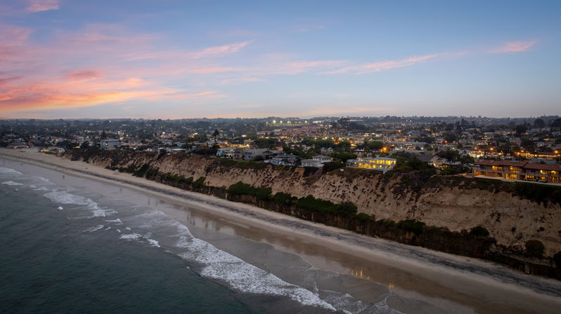 Sunset over a coastal bluff and sandy beach, gentle ocean waves lapping the shore with homes and twinkling town lights atop cliffs at dusk