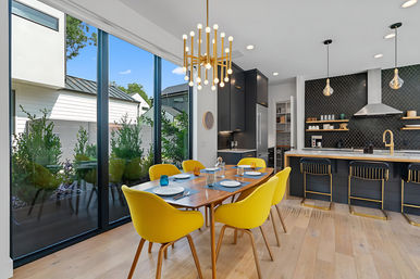 Bright open-concept modern dining room and kitchen in a contemporary suburban home — floor-to-ceiling windows, wooden table with six yellow chairs, black cabinets and island with gold accents, pendant lights, geometric tile backsplash and light oak floors.