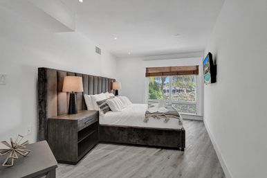 Bright modern bedroom with plush upholstered bed, bedside lamps and wood nightstand, folded towel swans on white linens, and a large window with bamboo blinds framing palm trees and a sunny tropical neighborhood view