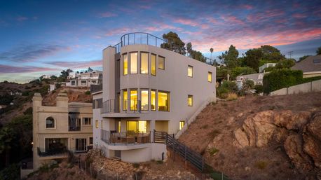 Modern multi-level coastal hillside home with curved corner windows and glowing interior lights, balconies overlooking a rocky bluff and neighboring houses under a pink-and-blue sunset sky.