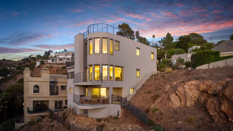 Modern multi-level coastal hillside home with curved corner windows and glowing interior lights, balconies overlooking a rocky bluff and neighboring houses under a pink-and-blue sunset sky.