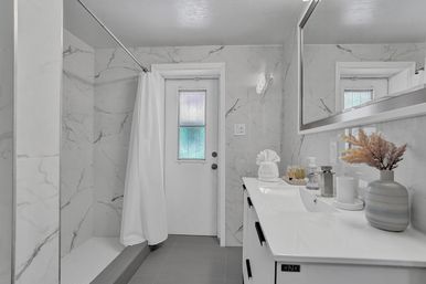 Spa-like modern bathroom with white marble-look walls, walk-in shower with curtain, frosted glass back door, sleek white vanity, large mirror, and a striped vase with dried grasses