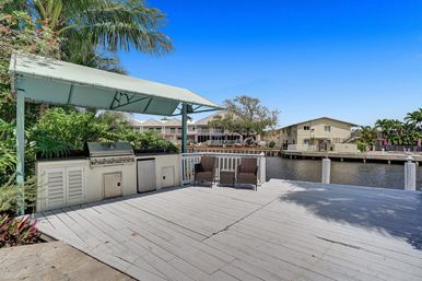 Canal-front wooden dock with a covered outdoor kitchen and stainless steel grill, two wicker chairs, tropical palms and waterfront condos under a bright blue sky