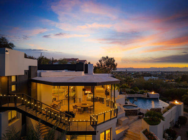 Modern luxury hillside home at desert sunset — glowing wraparound patio with string lights, outdoor dining, infinity pool and fire pits overlooking city lights and distant mountains.