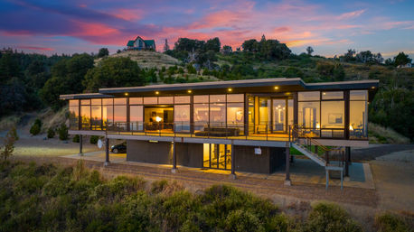 Sleek elevated modern glass house perched on a hillside at sunset, warm interior lights and wraparound deck offering panoramic views over a wooded valley
