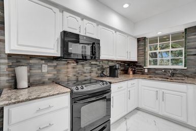 Bright modern kitchen with white cabinets, black microwave and range, granite-look countertops, gray stacked tile backsplash and a sink under a window with a leafy yard view.