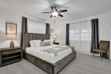Bright, tranquil contemporary master bedroom with upholstered king bed, white linens and textured throw, matching wood nightstands and lamps, ceiling fan, large windows with gray curtains and light wood floors.