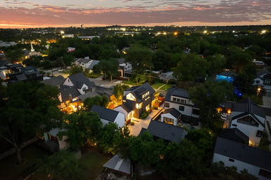 Aerial view of a tree-lined suburban neighborhood at dusk, modern homes with dark metal roofs and warm interior lights, illuminated driveways and a glowing backyard pool beneath a colorful sunset sky.