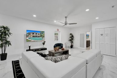 Bright modern white living room in a vacation-style home with a large white sectional sofa, marble tile floor, wall-mounted TV showing a pool and villa, wooden coffee table, ceiling fan, potted plants and recessed lighting.