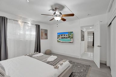 Sunlit modern master bedroom with wood-blade ceiling fan, wall-mounted TV displaying a backyard pool, gray curtains, neutral bedding with tassel throw, textured pouf and open doorway to a hallway.