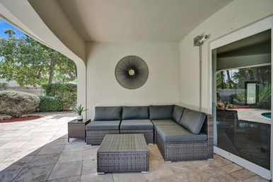 Cozy gray-cushioned rattan sectional and ottoman on a covered poolside patio nook with tiled floor, potted plant and decorative sunburst wall art.