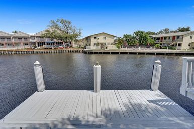 White wooden dock overlooking a calm canal with waterfront condos, palm trees, and a bright blue sky