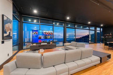 Modern open-plan living room with gray sectional sofa, hardwood floors, large floor-to-ceiling windows and balcony showcasing a panoramic ocean sunset, TV entertainment console and minimalist decor