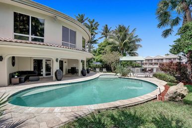 Luxury tropical backyard with curved kidney-shaped pool, two-story Mediterranean-style house, covered patio with lounge seating, palm trees and poolside chairs under a clear blue sky.