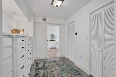 Bright white hallway in a modern home with built-in drawers and black pulls, teal patterned area rug, louvered closet doors, and a doorway opening to a sunlit bathroom with a round mirror.