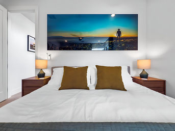 Cozy modern bedroom with crisp white bedding on a king-size bed, two brown accent pillows, matching wooden nightstands with lamps, and vibrant sunset landscape wall art featuring a silhouetted figure.