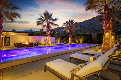 Twilight desert oasis with a glowing purple rectangular pool, palm trees, cushioned lounge chairs, a fire pit seating area and rugged mountain backdrop.