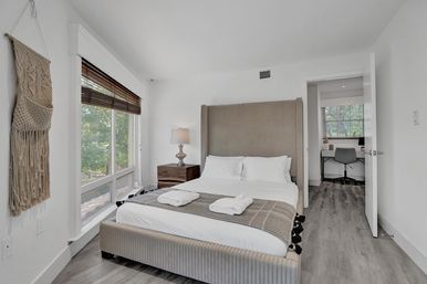 Bright modern minimalist bedroom with ribbed upholstered bed, white linens and folded towels, wooden nightstand and lamp, macramé wall hanging, large window with wooden blinds overlooking leafy trees, and an open door revealing a small home office.