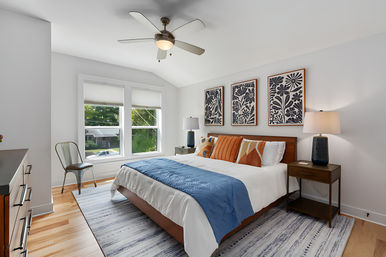 Bright contemporary master bedroom in a suburban home with vaulted ceiling and ceiling fan, large double window, wood bed with white linens, blue throw and orange accent pillows, three botanical prints above the headboard, matching nightstands and lamps, hardwood floors and striped area rug.