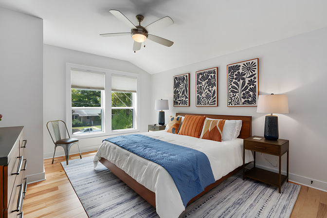 Bright contemporary master bedroom in a suburban home with vaulted ceiling and ceiling fan, large double window, wood bed with white linens, blue throw and orange accent pillows, three botanical prints above the headboard, matching nightstands and lamps, hardwood floors and striped area rug.