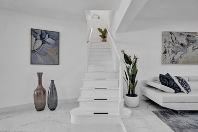 Contemporary white living room with marble floor and central staircase, abstract wall art, tall decorative vases, potted tropical plant, and white sectional sofa with blue pillows.