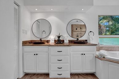 Modern coastal bathroom with white double vanity, stone countertop, round mirrors, vessel sinks and a tub by a palm-view window