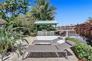 Tropical waterfront patio with two gray lounge chairs facing a white wooden deck, covered outdoor grill station under a green canopy, palm trees and lush landscaping, canal and waterfront townhomes in the background under a bright blue sky.