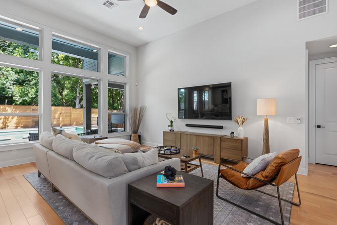 Sunlit modern living room with floor-to-ceiling windows overlooking a fenced backyard pool, neutral sectional, leather accent chair, wall-mounted TV and wooden media console on hardwood floors.