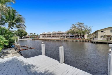 Sunny waterfront canal with a white wooden dock, palm trees, a boat lift, and waterside townhouses under a bright blue sky.