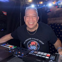 Smiling DJ in a nightclub booth leaning over a lit mixing console with colorful pad buttons and stage lights, wearing a black t-shirt and gold chain.