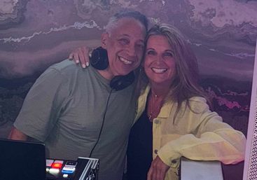 Two smiling people pose at a party—man wearing DJ headphones beside a woman in a light yellow jacket, leaning on DJ equipment in front of a purple marbled wall, warm indoor lounge vibe.