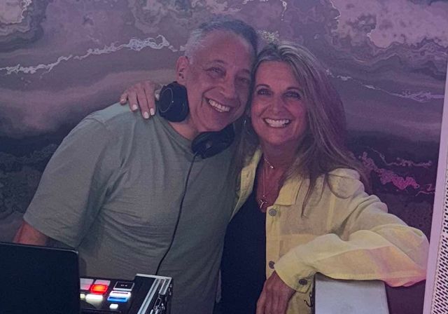 Two smiling people pose at a party—man wearing DJ headphones beside a woman in a light yellow jacket, leaning on DJ equipment in front of a purple marbled wall, warm indoor lounge vibe.