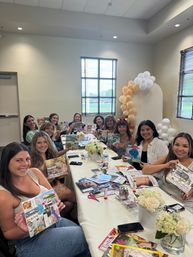 Smiling women at a vision-board workshop in a bright event room, gathered around a long table with magazines, scissors, glue sticks, floral centerpieces and a gold-and-white balloon arch.